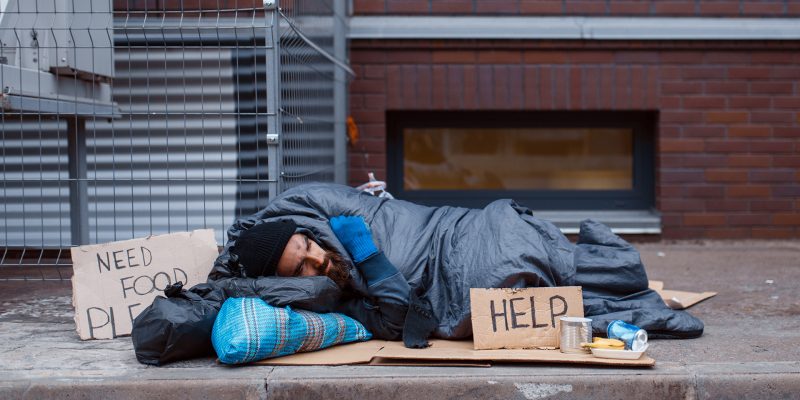 Bearded dirty homeless with help sign lies on city street. Poverty is a social problem, homelessness and loneliness, alcoholism and drunk addiction, urban lonely