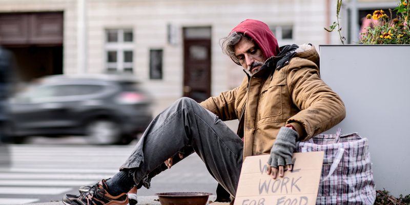 A homeless beggar man with a carboard sign sitting outdoors in city asking for money donation.